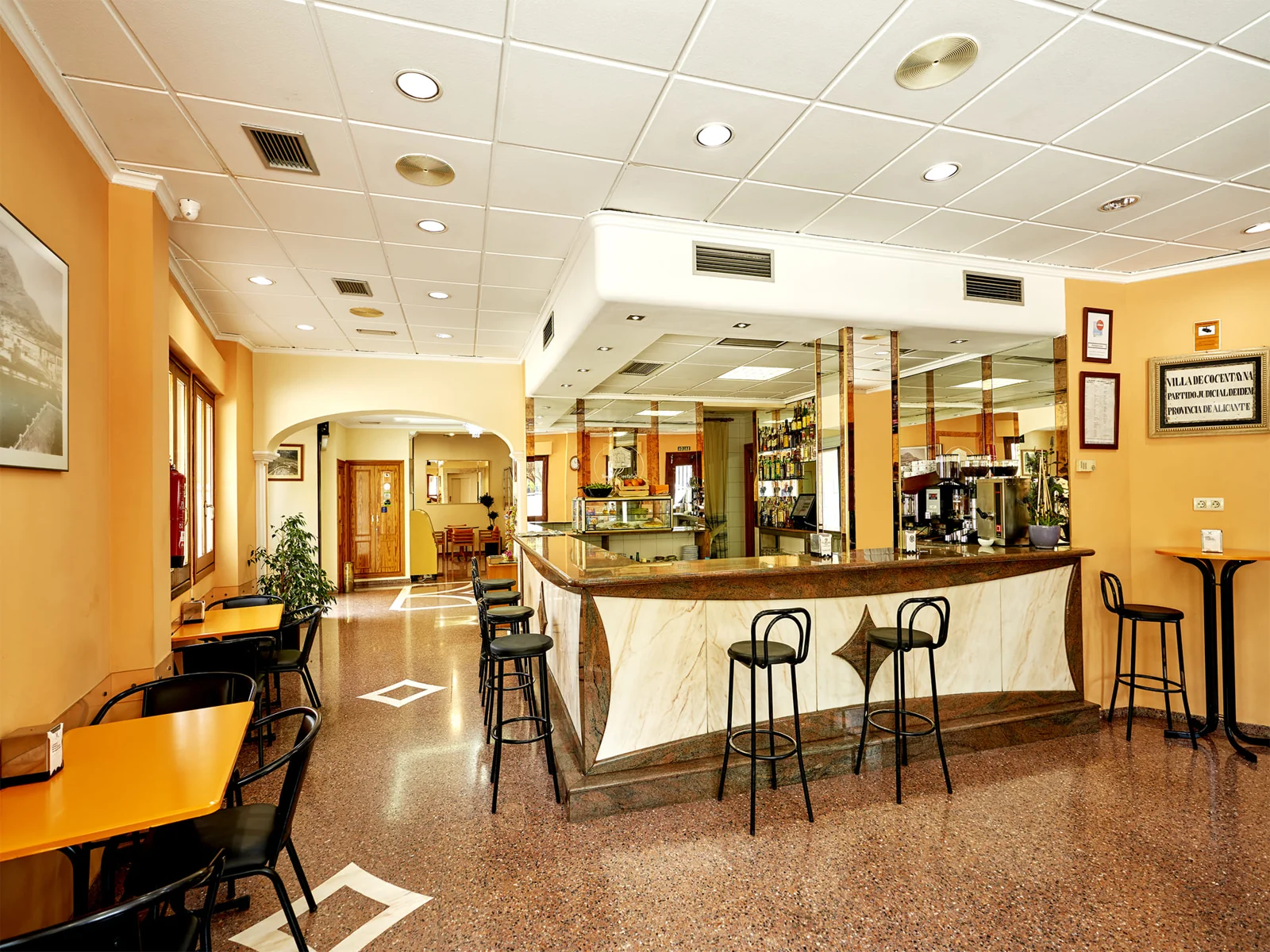 Interior real de la cafetería Hotel Anna, con barra de mármol, mesas y grandes ventanas